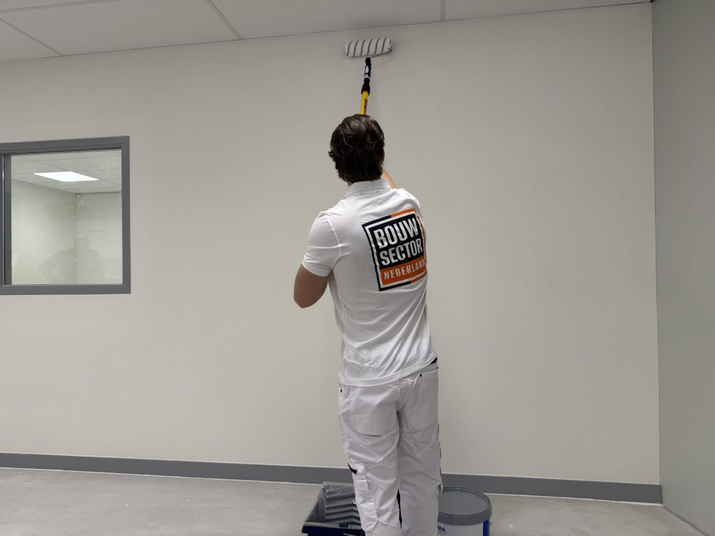 Schilder in witte werkkleding rolt met een verlengsteel een lichte muur tot aan het plafond