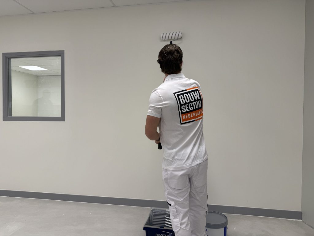 Schilder in wit werktenue rolt de bovenkant van een lichte wand onder het plafond, met verfbak en emmer