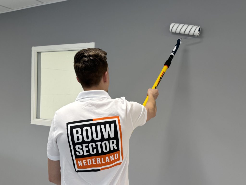 Schilder met het Bouwsector Nederland-logo rolt een grijze muur naast een binnenraam met wit kozijn